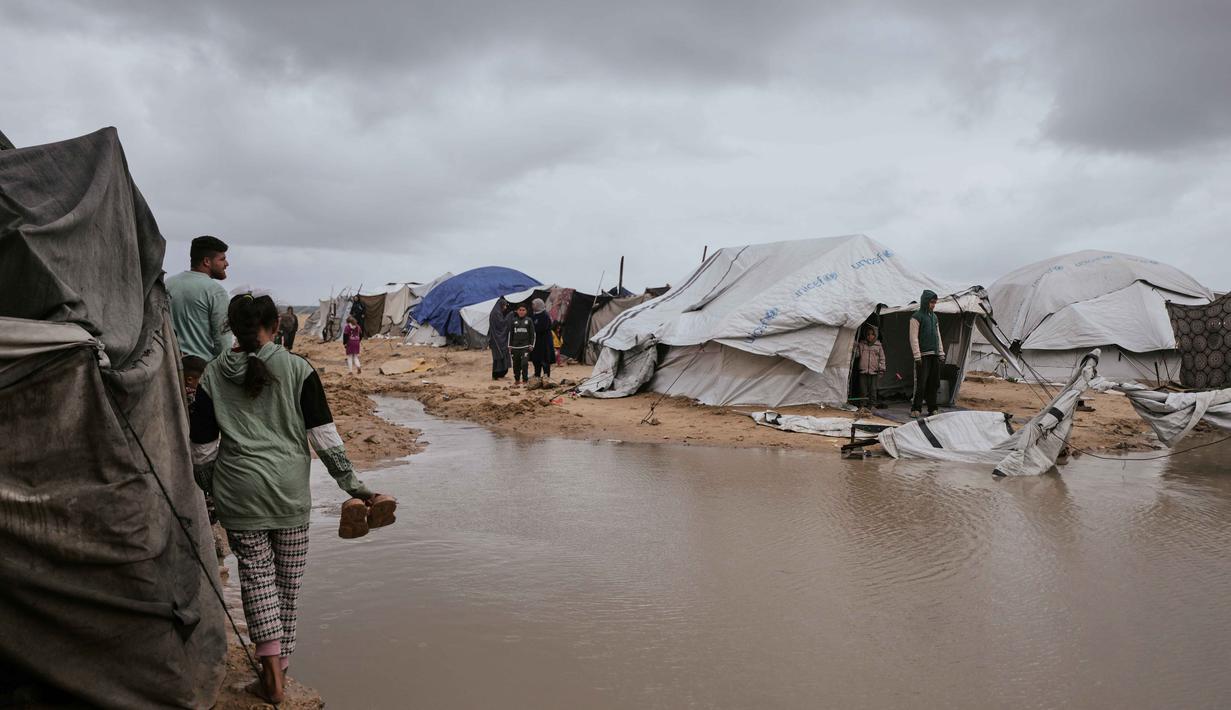 Genangan air menciptakan lingkungan yang tidak higienis. Tampak dalam foto, warga Palestina berjalan melewati area yang tergenang banjir di kamp tenda sementara setelah hujan lebat di Kota Gaza, Kamis, 26 Maret 2026 (AP Photo/Jehad Alshrafi)
