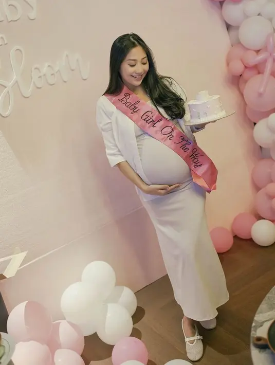 Saat baby shower, sambil memegang cake pink, Winona Willy mengenakan pakaian serba putih dari kemeja lengan pendek dipadukan inner dress panjang serta flat shoes ber jaringnya. [@nonawilly]