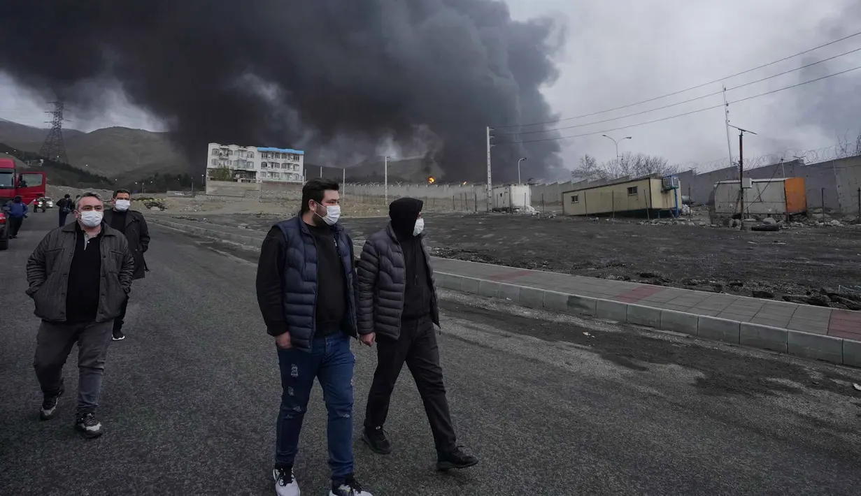 Media lokal Iran melaporkan kebakaran besar terjadi di Teheran dan mengonfirmasi bahwa kilang minyak dan fasilitas penyimpanan minyak terkena dua rudal serangan udara. Tampak dalam foto, empat pria berjalan berlatar kepulan asap tebal dari serangan gabungan AS-Israel terhadap fasilitas penyimpanan minyak pada Sabtu 7 Maret 2026 malam yang membubung ke langit di Teheran, Minggu, 8 Maret 2026. (AP Photo/Vahid Salemi)