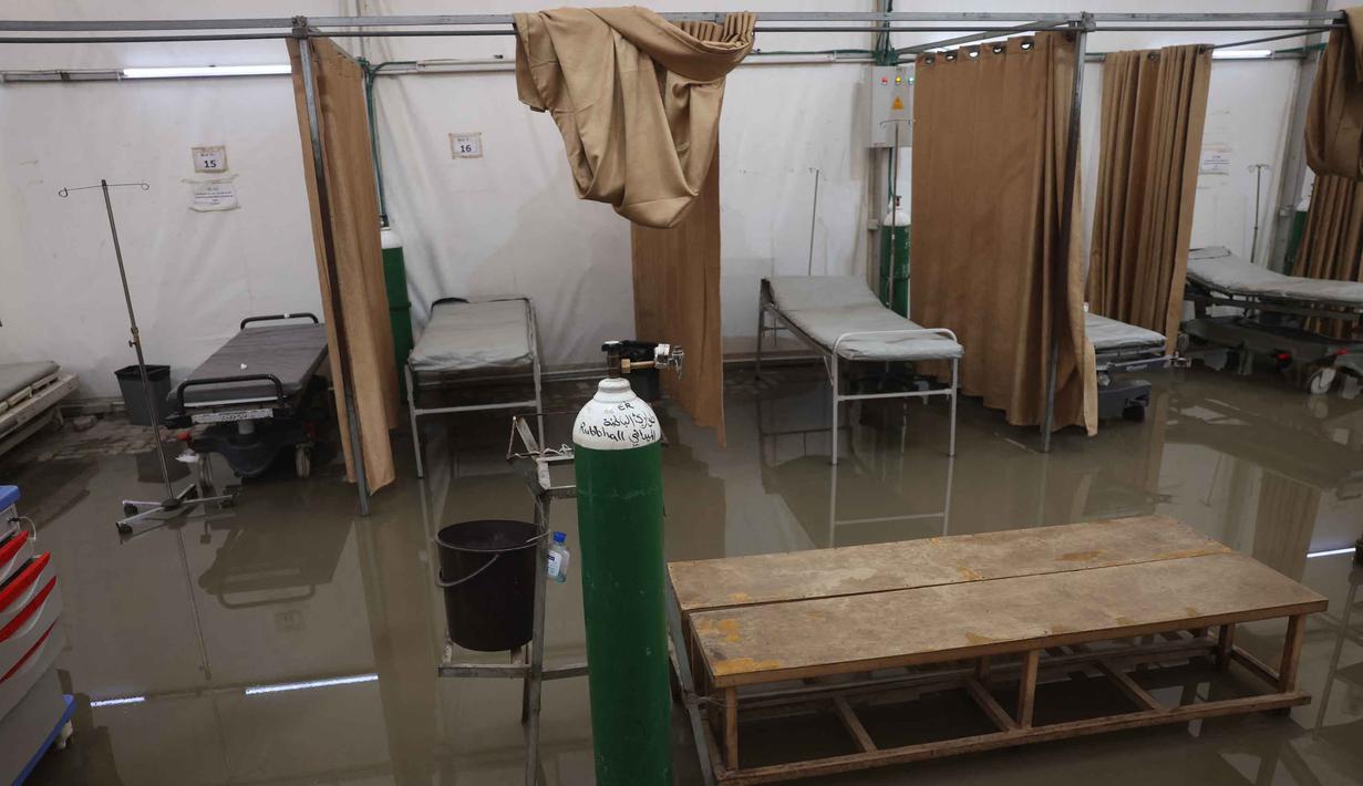 Kondisi ini memperparah krisis kemanusiaan di wilayah tersebut. Tampak dalam foto, ranjang-ranjang kosong di dalam tenda darurat milik Rumah Sakit Nasser, yang berfungsi sebagai tempat penerimaan pasien rawat jalan dan departemen gawat darurat, setelah hujan lebat di Khan Yunis di Jalur Gaza selatan pada 26 Maret 2026. (Bashar Taleb/AFP)