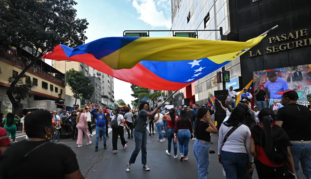 Mereka meneriakkan slogan anti-imperialisme dan menyatakan bahwa Venezuela bukan negara yang akan menyerah. Tampak dalam foto, seorang pendukung mantan Presiden Venezuela, Nicolas Maduro, membawa bendera nasional saat mengikuti aksi unjuk rasa di Caracas pada Minggu 4 Januari 2026. (Juan BARRETO/AFP)
