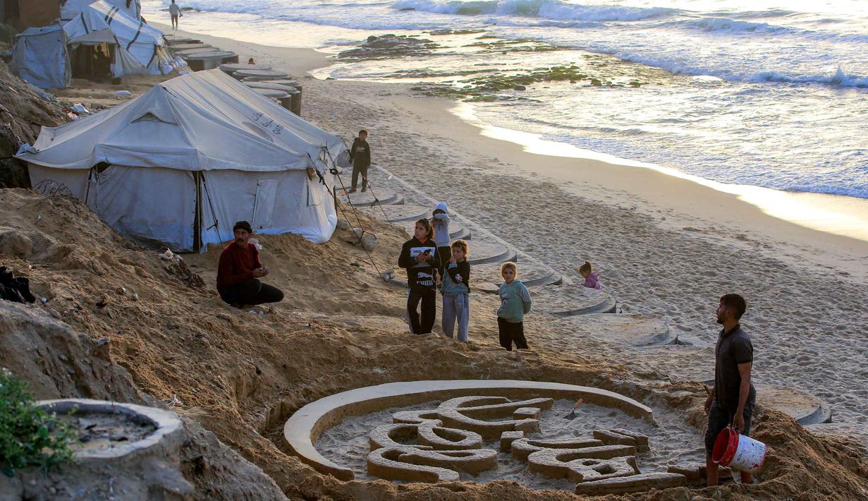 Meski bersifat sementara karena sering terhapus oleh pasang air laut di malam hari, namun Yazeed Abu Jarad bersama timnya secara rutin kembali setiap pagi untuk membuat karya baru sebagai simbol harapan. Tampak dalam foto, warga membantu seniman Palestina Yazeed Abu Jarad, saat ia membuat patung pasir dengan pesan ucapan "Selamat Datang, Ramadan" di sepanjang pantai di Khan Younis, selatan Gaza pada 17 Februari 2026. (Bashar Taleb/AFP)