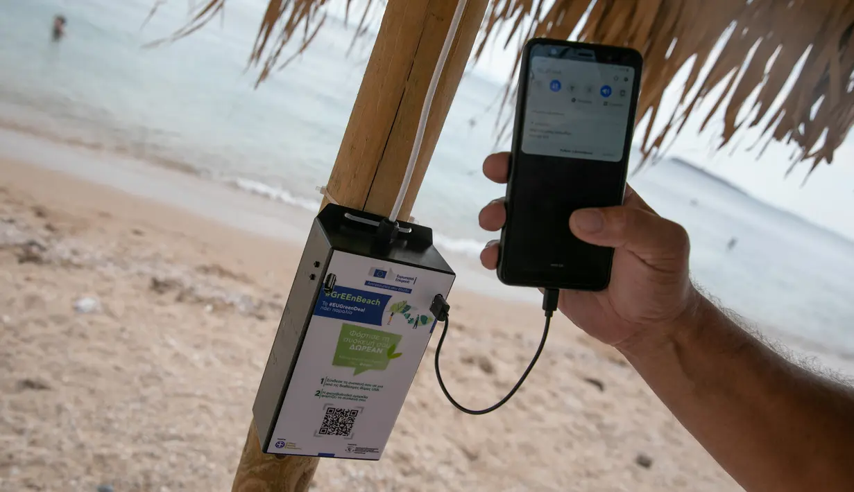 Ponsel diisi dayanya menggunakan sistem panel surya dan port USB di salah satu payung pantai dekat Balai Kota Vari-Voula-Vouliagmeni, Yunani, Selasa (21/7/2020). Para pengunjung pantai dapat berkontribusi terhadap upaya penghematan energi menggunakan tenaga surya. (Xinhua/Lefteris Partsalis)