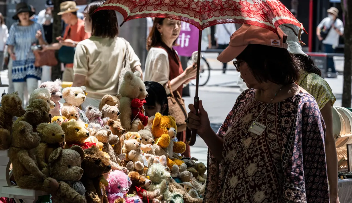 Seorang perempuan (kanan) menggunakan payung untuk berlindung dari teriknya sinar matahari saat melihat-lihat boneka beruang yang dijual di pasar barang antik di pusat kota Tokyo, Jepang, Senin (17/7/2023). (Richard A. Brooks / AFP)