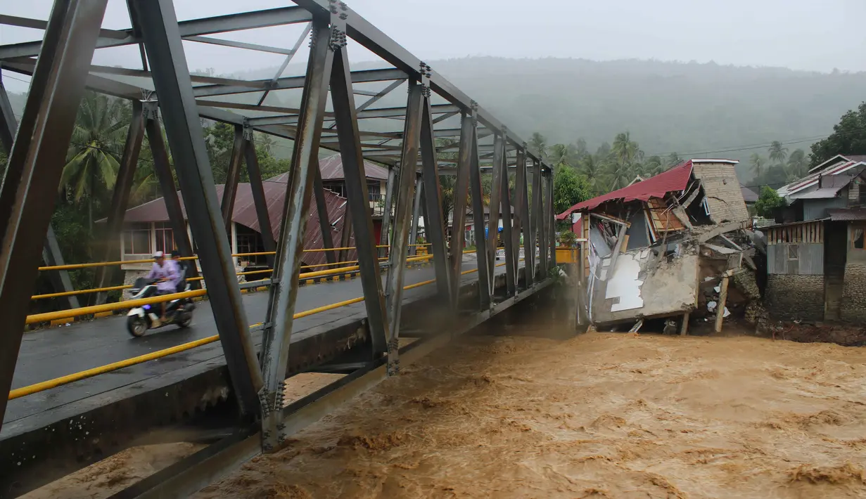 Sedikitnya 62 nyawa melayang, puluhan ribu warga terpaksa mengungsi. Beberapa akses vital di tiga provinsi utama: Aceh, Sumatera Utara, dan Sumatera Barat putus. Tampak dalam foto, pengendara melintasi jembatan melewati bangunan yang rusak akibat banjir di Tanah Datar, Sumatera Barat, Jumat 28 November 2025. (AP Photo/Ali Nayaka)