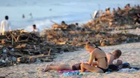 Turis asing bersantai dekat sampah yang terdampar akibat cuaca buruk di Pantai Kuta, Bali, Jumat (15/2). Hujan deras disertai angin kencang yang melanda Bali berdampak pada arus laut yang terus membawa sampah dari daerah lain. (SONNY TUMBELAKA/AFP)