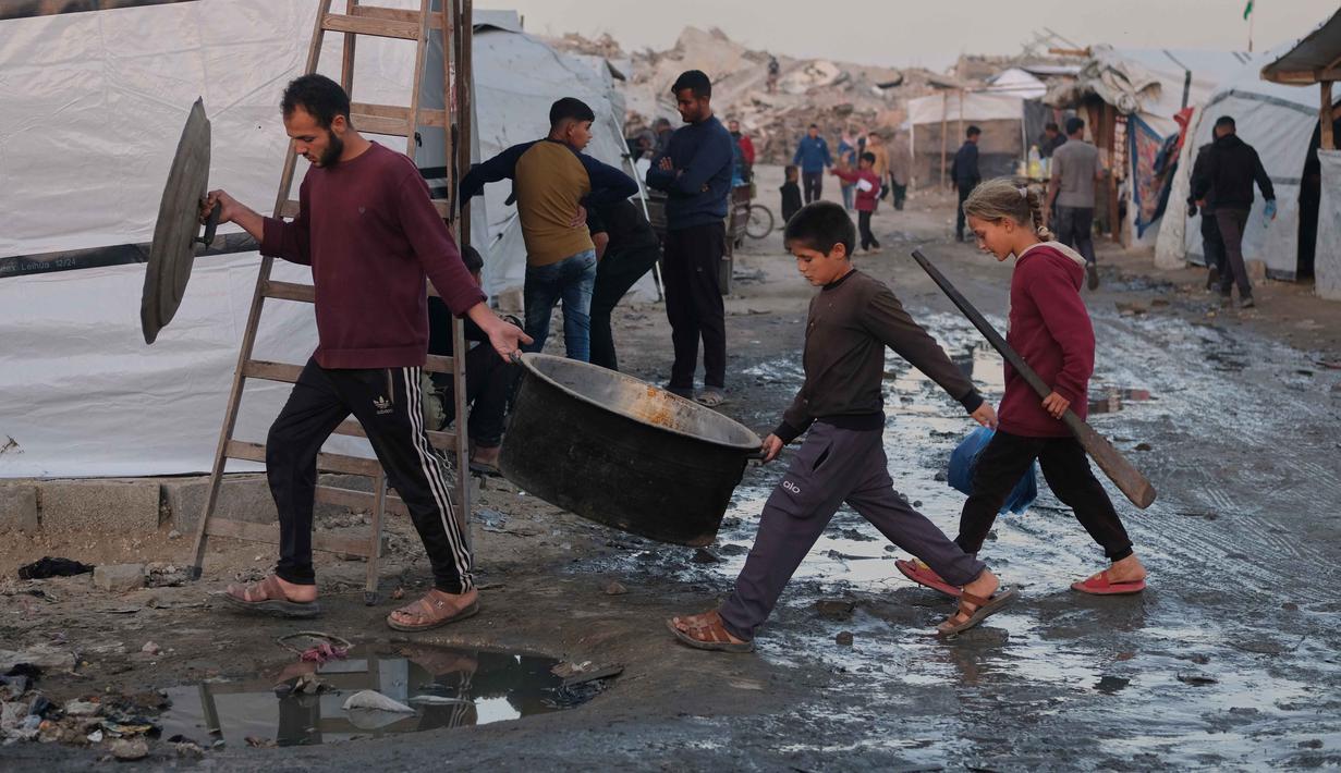 Situasi ini menghambat upaya pemulihan. Tampak dalam foto, pengungsi Palestina berjalan melalui jalan-jalan berlumpur sementara seorang pria dan anak-anak membawa peralatan masak di sebuah kamp di Jabalia, di bagian utara Jalur Gaza, Minggu 7 Desember 2025. (AP Photo/Jehad Alshrafi)
