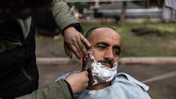 Khaled al-Hamo sedang dipotong rambut oleh putranya, Mohammed al-Hamo, di depan tenda mereka di kamp darurat di kota Antakya pada 19 Februari 2023. Hamo menawarkan jasa memotong rambut di ruang terbuka untuk keluarganya dan mereka yang kehilangan tempat tinggal akibat gempa dahsyat yang menimpa Turki dan Suriah pada 6 Februari 2023. (Sameer Al-DOUMY / AFP)