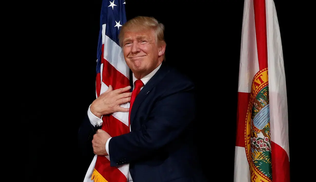 Capres AS dari Partai Republik, Donald Trump memeluk bendera AS saat melakukan kampanye menyapa pendukungnya di Tampa, Florida, AS (24/10). Trump akan bertarung dengan Hillary Clinton memperebutkan kursi di Gedung Putih. (REUTERS/Jonathan Ernst)