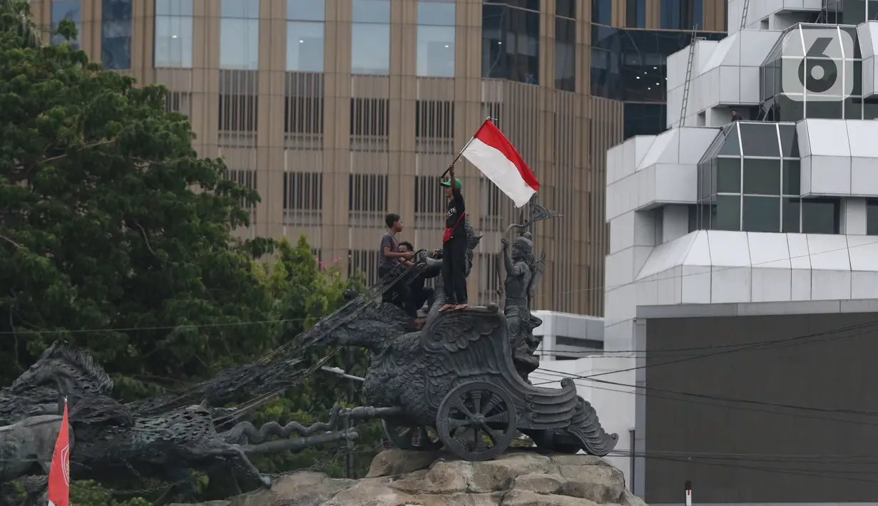 Massa aksi demo tolak omnibus law UU Cipta Kerja naik ke atas patung arjuna wiwaha mengibarkan bendera merah putih di Jalan Medan Merdeka Barat, Jakarta Pusat, Selasa (20/10/2020). Aksi mereka lantas disambut meriah oleh para massa yang masih bertahan. (Liputan6.com/Herman Zakharia)