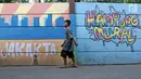 Seorang anak kecil berjalan di sisi tembok yang terdapat lukisan mural di kawasan Kramat Jati, Jakarta, Rabu (4/4). Mural yang ada di tempat ini mencerminkan pesan-pesan sosial positif kepada masyarakat. (Liputan6.com/Herman Zakharia)