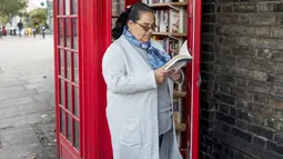 Seorang wanita membaca buku di dekat kotak telepon merah yang telah berubah menjadi perpustakaan pertukaran buku di Lewisham Way, London (21/10). Kotak telepon ini diubah menjadi perpustakaan, kios, hingga tempat reparasi ponsel. (AFP Photo/Tolga Akmen)