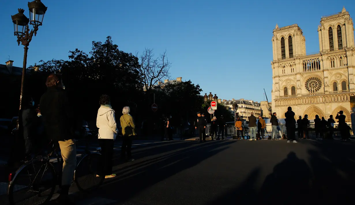 Orang-orang berhenti di depan Katedral Notre-Dame yang kembali membunyikan lonceng utamanya di Paris, Rabu (15/4/2020). Lonceng utama katedral kembali dibunyikan setelah satu tahun mengalami kebakaran yang sampai mengejutkan seluruh warga dunia. (AP/Christophe Ena)
