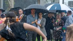 Pangeran Harry dan Meghan Markle berlindung di bawah payung menyaksikan tarian aborigin saat piknik di Victoria Park, Dubbo, Australia, Rabu (17/10). Keduanya diagendakan tur ke Australia dan Pasifik Selatan selama 16 hari. (Ian Vogler/Pool via AP)