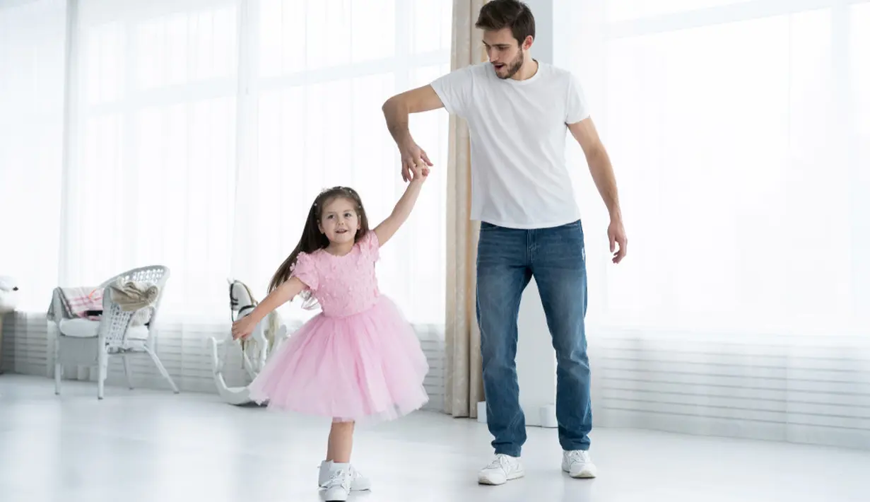 Seorang ayah sedang menari dengan anaknya di dalam rumah. Terlihat mereka berdua bepegangan tangan dan memeragakan tarian. Sebuah potret kedekatan anak dan ayah yang sangat manis sekali ya. (OPOLJA/ Shutterstock.com)
