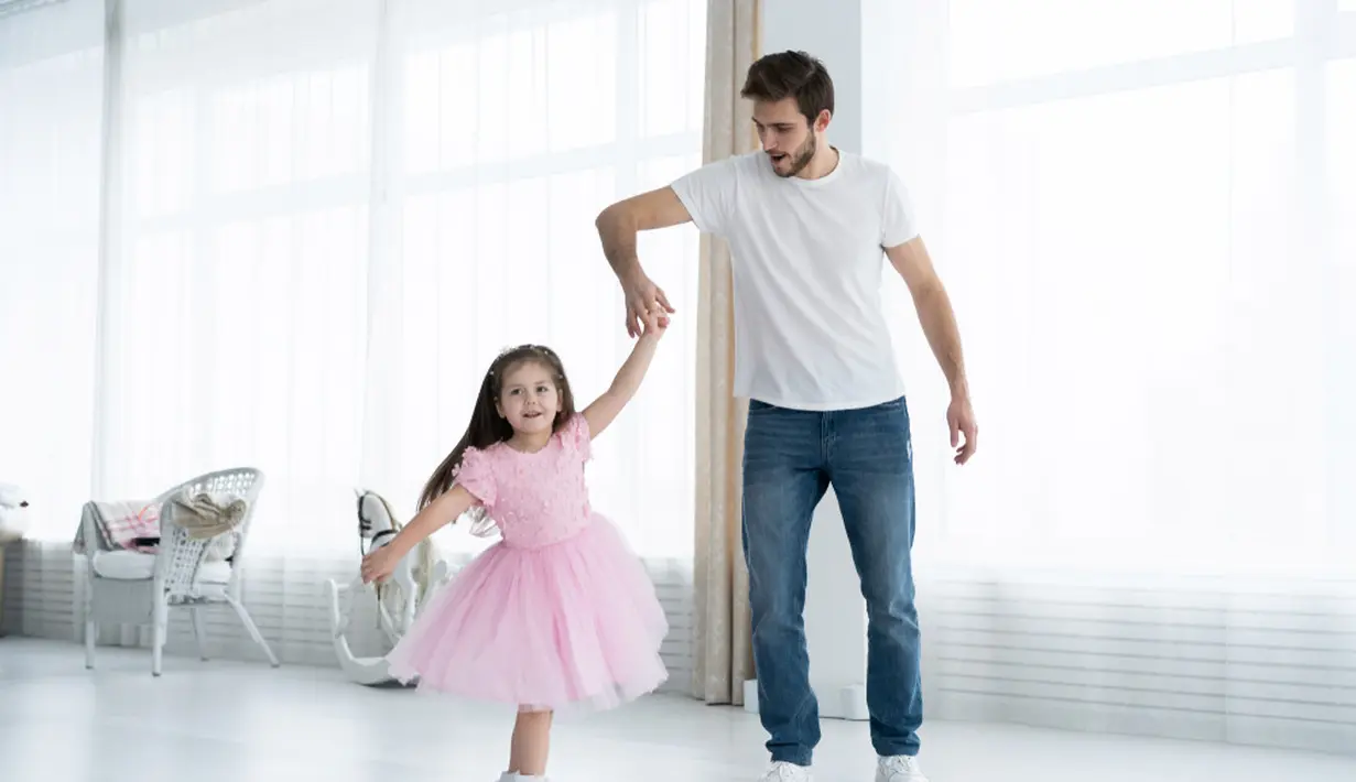 Seorang ayah sedang menari dengan anaknya di dalam rumah. Terlihat mereka berdua bepegangan tangan dan memeragakan tarian. Sebuah potret kedekatan anak dan ayah yang sangat manis sekali ya. (OPOLJA/ Shutterstock.com)