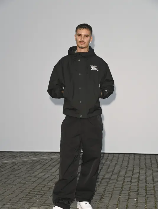 Romeo Beckham tampil kasual dengan jaket hoodie teknikal hitam berlogo kuda Burberry, celana lebar hitam, dan sneakers putih [dok. Burberry]