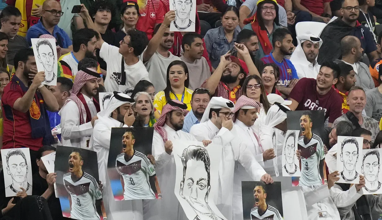 Penonton memegang foto Mesut Ozil di tribun dan menutup mulut mereka selama pertandingan Spanyol vs Jerman pada laga Grup E Piala Dunia 2022  di Stadion Al Bayt, Senin (28/11/2022) dini hari WIB. Mesut Ozil, yang merupakan keturunan imigran Turki kelahiran Jerman, merasa mengalami diskriminasi setelah dijadikan sebagai kambing hitam atas keluarnya Jerman dari Piala Dunia 2018 Rusia saat itu. (AP Photo/Matthias Schrader)