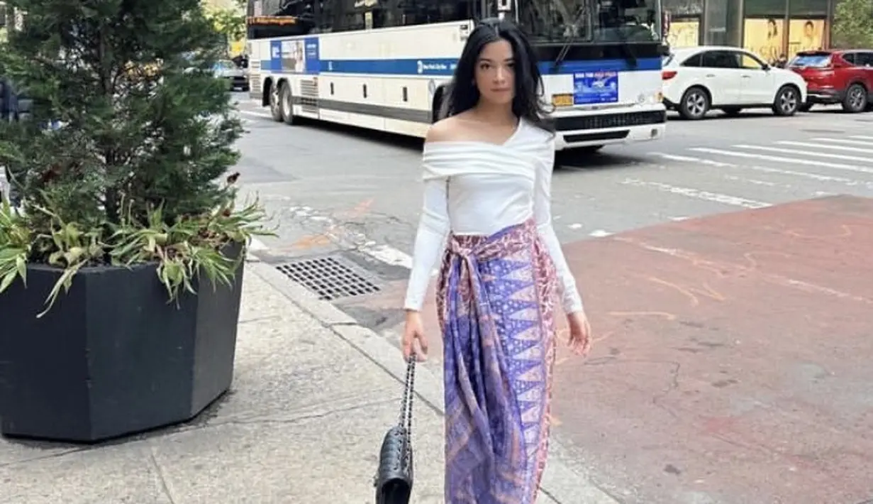 Saat berada di Amerika, Yoriko Angeline mengenakan atasan putih lengan panjang model off shoulder dipadukan kain lilit batik warna pink keunguan, serta sandal putihnya. [@yorikooangln_]