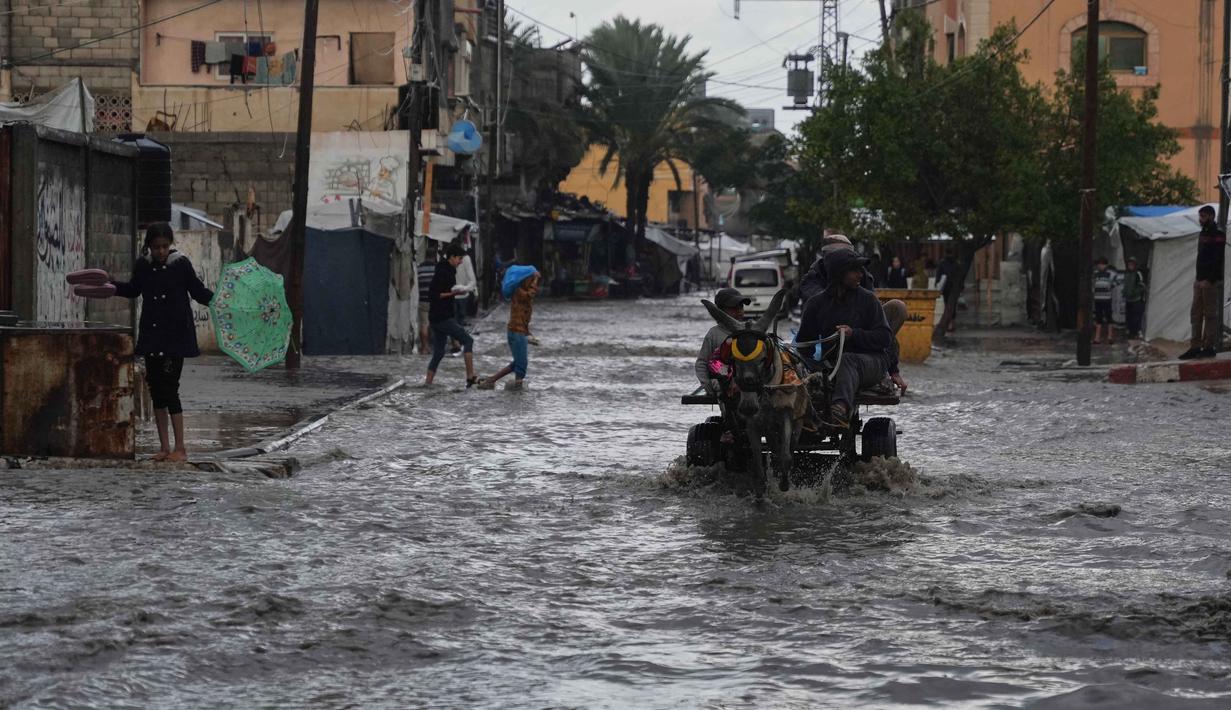 Serangan Israel juga memaksa sebagian besar warga untuk mengungsi ke tenda-tenda yang tidak memberikan perlindungan maksimal. Tampak dalam foto, warga Palestina berjalan di jalanan yang terendam banjir setelah hujan deras di Deir al-Balah, Jalur Gaza tengah, Selasa 25 November 2025. (AP Photo/Abdel Kareem Hana)