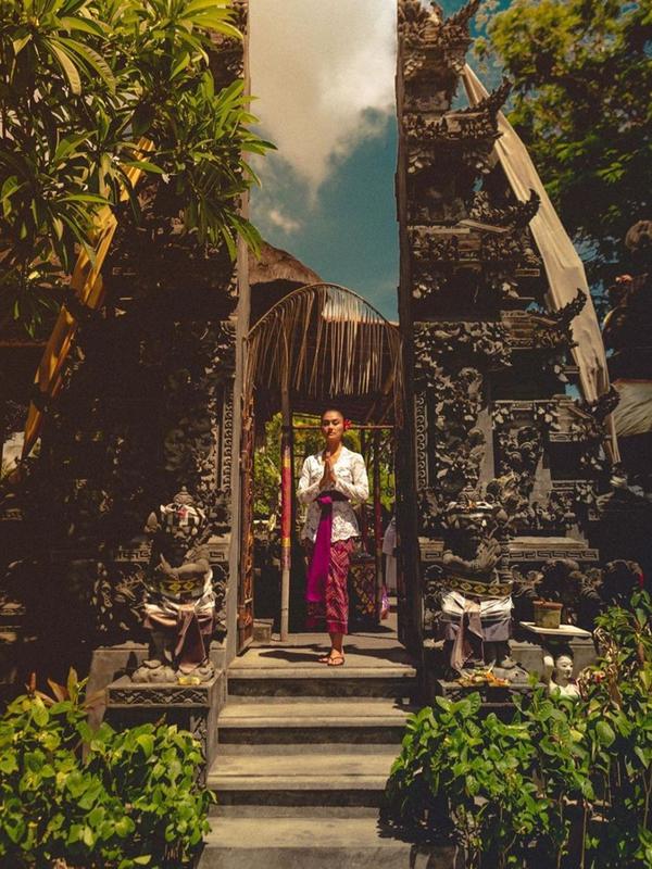 Mempelajari budaya Bali saat di sana, pesona Agnez Mo ketika mengenakan busana adat Bali mencuri perhatian saat berpose di gapura. Ia mengenakan kebaya renda warna putih yang dipadukan dengan kain warna ungu. (Liputan6.com/IG/@agnezmo)