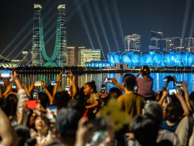 Orang-orang berkumpul di kawasan pejalan kaki Sungai Qiantang untuk menyaksikan pertunjukan cahaya di Hangzhou Olympic Sports Centre Stadium (kanan) menjelang Asian Games 2023 di Hangzhou, Provinsi Zhejiang, China, Selasa (19/9/2023). Hangzhou Olympic Sports Centre Stadium merupakan lokasi pembukaan Asian Games 2023. (Philip FONG/AFP)