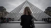 Seorang perempuan berdiri di depan Pyramid, pintu masuk utama menuju museum Louvre, di Paris, Rabu (4/3/2020). Louvre di Paris, museum yang paling banyak dikunjungi di dunia, kembali dibuka setelah pegawai yang khawatir dengan penularan virus corona setuju kembali masuk kerja. (Philippe LOPEZ/AFP)
