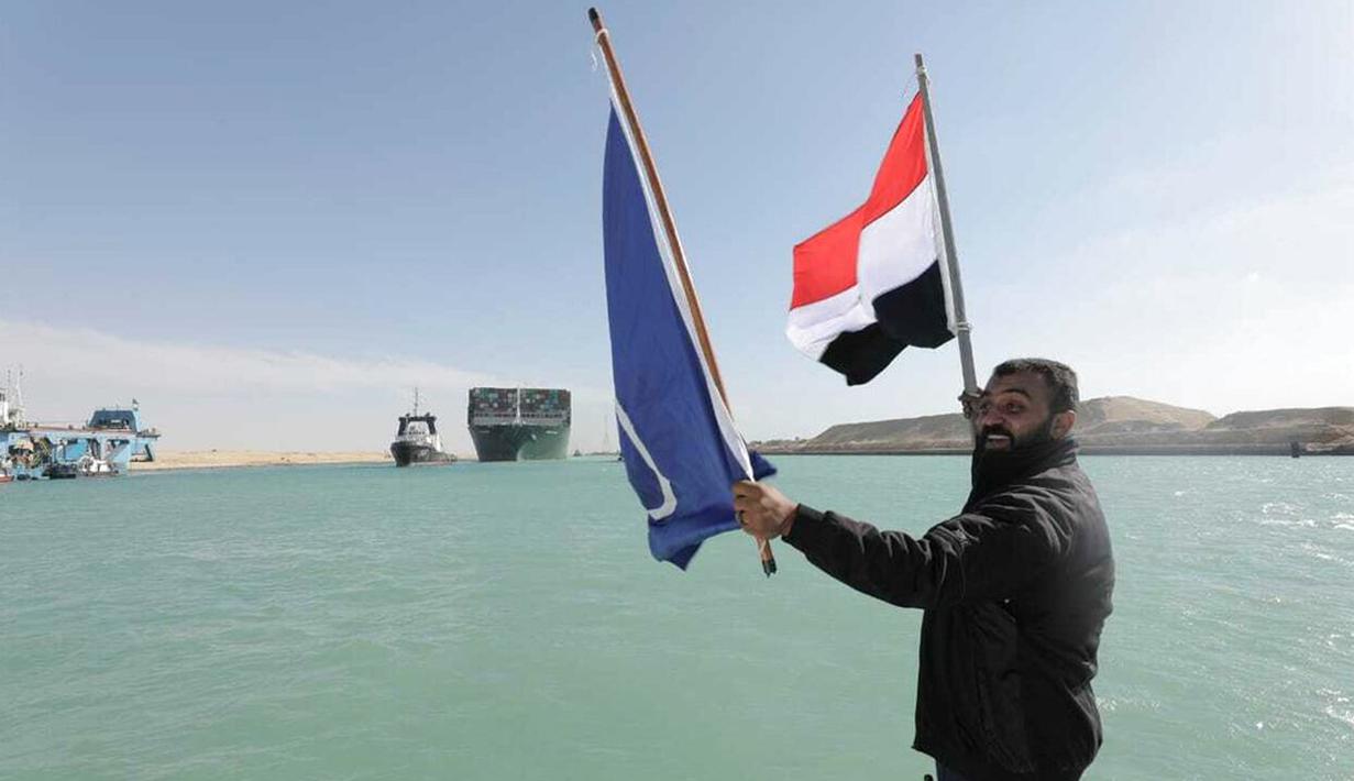 Seorang pria mengibarkan bendera saat kapal kargo Ever Given ditemani kapal tunda melaju di Terusan Suez, Mesir, Senin (29/3/2021). Para ahli percaya efek lanjutan pada pengiriman global bisa memakan waktu berminggu-minggu atau bahkan berbulan-bulan untuk diselesaikan. (Suez Canal Authority via AP)