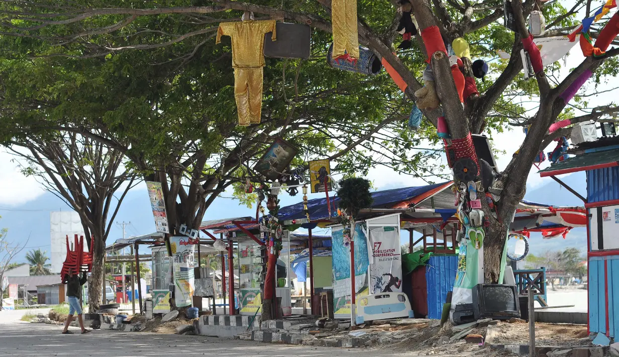 Seorang pria membawa kursi melewati pohon yang berhias barang-barang untuk memperingati gempa dan tsunami Palu, Rabu (3/4). Kondisi Palu mulai membaik setelah bencana gempa dan tsunami melanda melanda enam bulan lalu. (OLAGONDRONK/AFP)