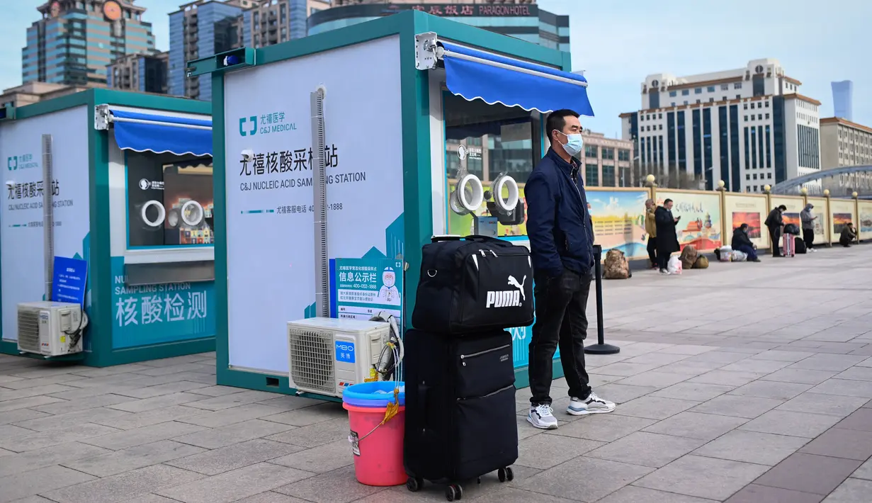 Seorang pria membawa tas menunggu kereta di sebelah bilik pengujian asam nukleat di luar pintu masuk stasiun kereta api Beijing di ibu kota China (7/1/2023). Ketika migrasi tahunan dimulai dengan orang-orang yang kembali ke kampung halaman mereka untuk perayaan Tahun Baru Imlek. (AFP/Wang Zhao)