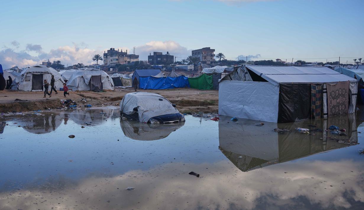 Banjir juga memicu risiko penyakit menular akibat sistem drainase yang hancur akibat serangan Israel sejak Oktober 2023 lalu. Tampak dalam foto, anak-anak Palestina berlari melewati sebuah mobil yang sebagian terendam di daerah yang tergenang banjir setelah hujan lebat di sebuah kamp pengungsian sementara, Zawaida, Jalur Gaza tengah, Rabu 17 Desember 2025. (AP Photo/Abdel Kareem Hana)