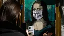 Seorang perempuan mengambil foto poster seniman Italia Salvatore Benintende yang menggambarkan Mona Lisa karya Leonardo da Vinci mengenakan masker dan memegang smartphone yang bertuliskan "Mobile World Virus" di sebuah jalan di kota Barcelona, Spanyol pada Selasa (18/2/2020). (PAU BARRENA/AFP)
