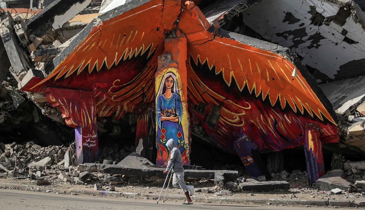 Langkah ini dianggap penting untuk sepenuhnya mengimplementasikan kesepakatan gencatan senjata yang rapuh antara Israel dan Hamas dan bergerak menuju fase kedua rencana perdamaian. Tampak dalam foto, seorang pria menggunakan kruk melewati mural di sisi-sisi bangunan yang hancur di kamp pengungsi Maghazi di luar Deir al-Balah pada Senin 8 Desember 2025. (EYAD BABA/AFP)