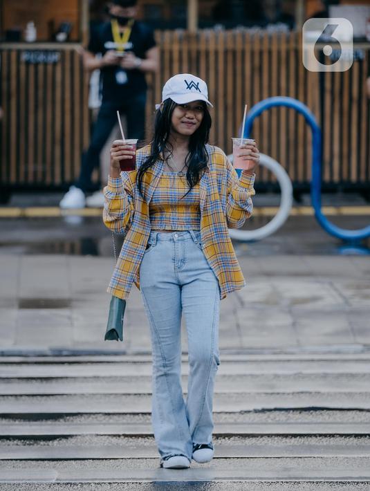 Seorang remaja wanita membawa minuman berjalan melintasi zebra cross di Taman Sudirman, Jakarta, Rabu (6/7/2022). Tempat ini viral karena jadi ajang adu fashion anak Citayam, Bekasi hingga Bojong Gede. Mereka mengenakan kemeja flanel oversize, celana model 90-an seperti boot cut atau cutbray, sneakers klasik, dan tentu saja topi. (Liputan6.com/Faizal Fanani)