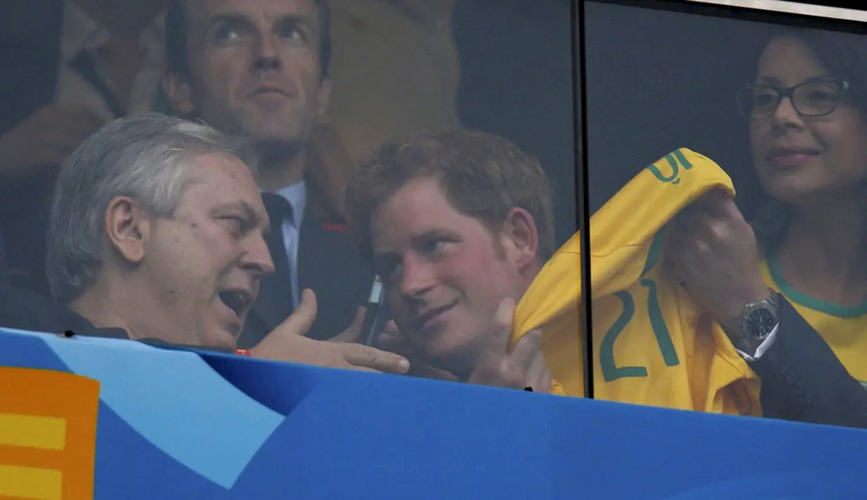 Dari balik kaca pelindung, Pangeran Harry terlihat memamerkan kaos Timnas Brasil kepada salah satu penonton yang ikut menyaksikan laga Brasil vs Kamerun di Stadion Nasional Brasil, (24/6/2014). (REUTERS/Ueslei Marcelino)