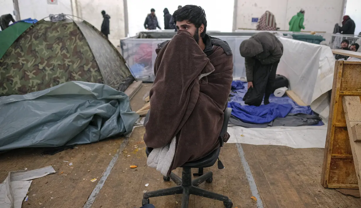 Seorang migran yang membungkus tubuh dengan selimut duduk di kursi di tempat penampungan sementara di kamp Lipa, barat daya Bosnia, Sabtu (26/12/2020). Ratusan migran terdampar di tenda-tenda kumuh dan terbakar di Bosnia, di tengah hujan salju yang lebat dan suhu membeku. (AP Photo/Kemal Softic)