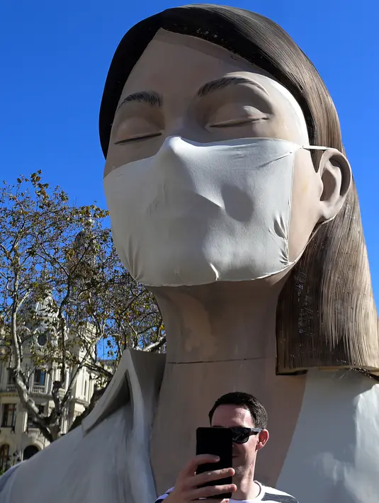 Seorang pria berselfie di depan patung raksasa mengenakan masker yang akan ditampilkan di festival Las Fallas atau festival api di Valencia, Spanyol, Rabu (11/3/2020). Festival Fallas yang akan berlangsung pada 13 Maret telah dibatalkan karena wabah coronavirus. (AFP/Jose Jordan)