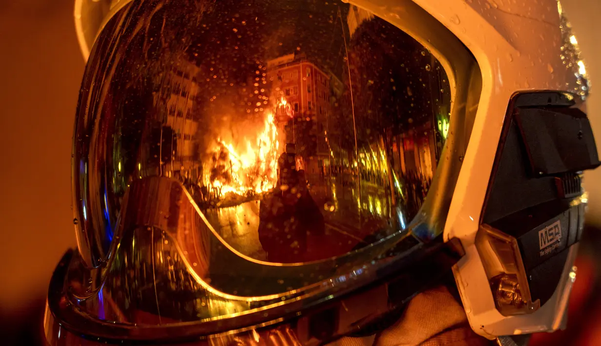 Falla terbakar saat tercermin dalam helm pemadam kebakaran selama festival Fallas tradisional di Valencia (19/3/2022). Fallas adalah struktur raksasa yang terbuat dari karton yang menggambarkan peristiwa dan kepribadian terkini di mana tokoh individu atau Ninot ditempatkan. (AP Photo/Alberto Saiz)