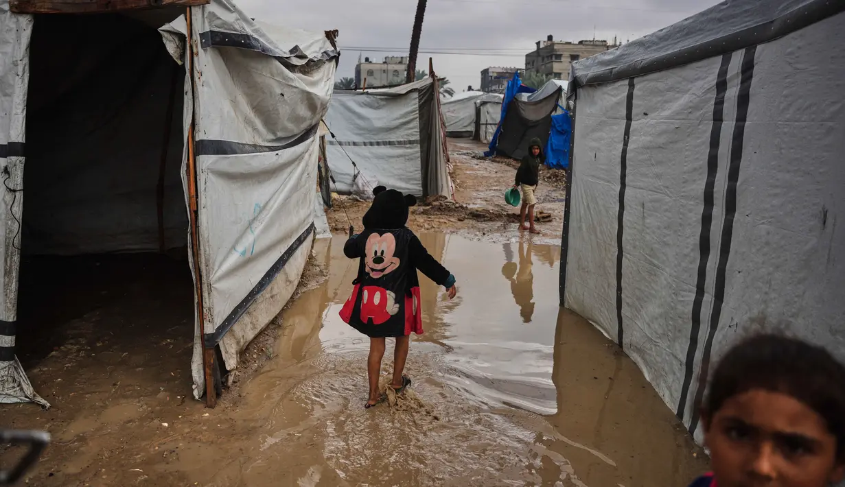 Situasi ini memperburuk kehidupan warga Gaza yang tinggal di lokasi penampungan. Tampak dalam foto, warga Palestina berjalan melewati tenda-tenda sementara yang terendam banjir setelah hujan deras di Deir al-Balah, Jalur Gaza tengah, Selasa 25 November 2025. (AP Photo/Abdel Kareem Hana)