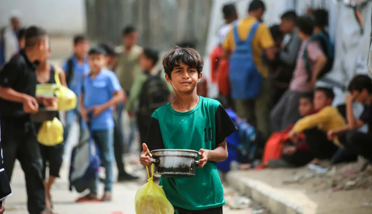 Seorang anak laki-laki Palestina yang mengungsi membawa sepanci makanan yang dikumpulkan dari dapur amal ke tempat penampungan keluarganya, di kamp pengungsi Bureij, Jalur Gaza Tengah pada Senin 18 Mei 2026. (Eyad Baba/AFP)