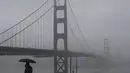 Pejalan kaki berdiri di bawah payung di jalan setapak di depan Jembatan Golden Gate di San Francisco, Rabu (11/1/2023). California yang dilanda badai berjuang untuk membersihkan dan memperbaiki kerusakan yang meluas. (AP Photo/Jeff Chiu)