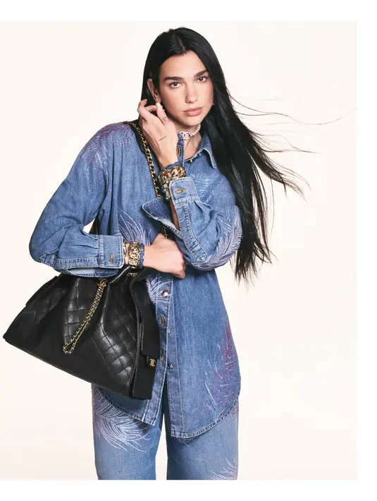 Kemeja denim dengan kancing terbuka memberikan kesan effortless chic, sedangkan aksesori seperti gelang rantai emas dan choker menambah kesan edgy. Tas CHANEL 25 berwarna hitam dengan rantai emas melengkapi tampilan, menunjukkan keseimbangan antara gaya kasual dan mewah. [Dok/CHANEL].