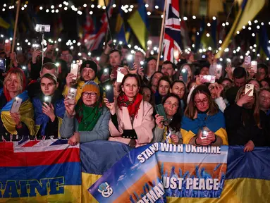 Orang-orang memegang plakat, bendera nasional Ukraina, dan menyalakan lampu ponsel mereka saat berkumpul untuk mendukung Ukraina yang menandai tahun ketiga invasi Rusia di Trafalgar Square, pusat kota London, pada 24 Februari 2025. (HENRY NICHOLLS/AFP)