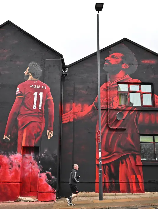Seseorang berolahraga melewati mural seniman John Culshaw yang menggambarkan pesepak bola Mohamed Salah di fasad bangunan dekat Stadion Anfield, Liverpool, Rabu (30/3/2022). Salah tidak diragukan lagi, adalah salah satu pemain terhebat yang pernah mengenakan seragam Liverpool. (Paul ELLIS / AFP)