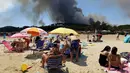 Wisatawan berjemur sambil menyaksikan asap hitam yang membumbung akibat kebakaran hutan dari pinggir pantai Lavandou, Rabu (26/7). Kebakaran menghanguskan hamparan hutan di Prancis selatan dan di Pulau Corsica dalam tiga hari terakhir. (AP/Claude Paris)