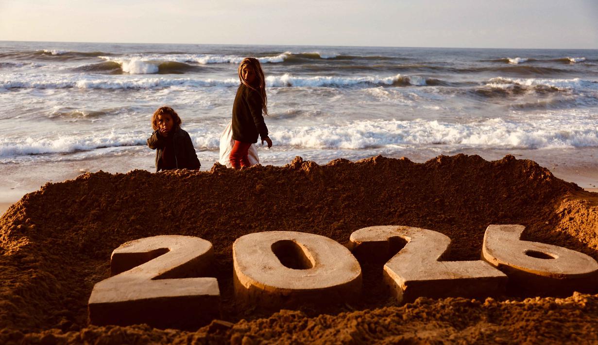 Anak-anak berjalan di belakang patung pasir karya seniman Yazed Abo Jarad yang menggambarkan angka 2026 atau tahun mendatang di Deir El-Balah, Jalur Gaza tengah pada 30 Desember 2025. Para pengungsi Palestina yang berada di Deir El-Balah, Jalur Gaza Tengah, bersiap menyambut pergantian tahun. (Bashar Taleb/AFP)