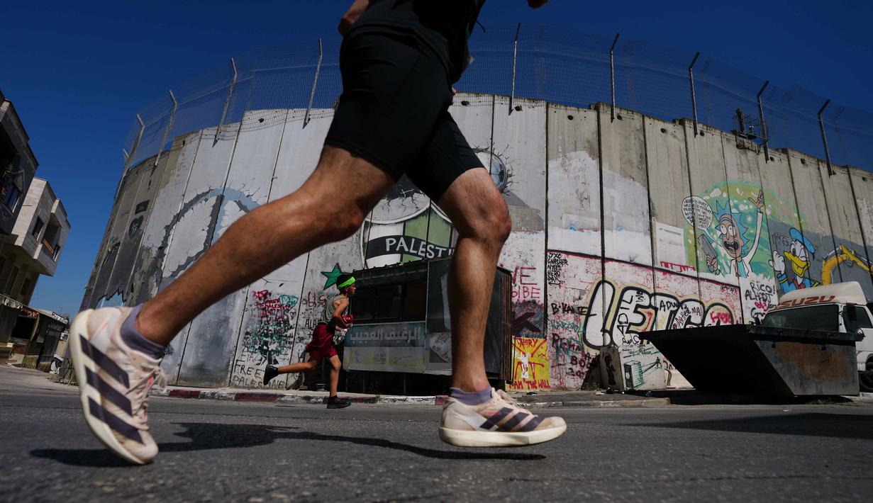 Sebagai informasi, edisi tahun ini, Maraton Internasional Palestina, diikuti oleh lebih dari 13.000 pelari, termasuk sekitar 1.000 peserta internasional dari 75 negara. Tampak dalam foto, para pelari melewati tembok pemisah Israel saat mereka berkompetisi dalam Maraton Palestina di kota Betlehem, Tepi Barat, Jumat 8 Mei 2026. (AP Photo/Mahmoud Illean)