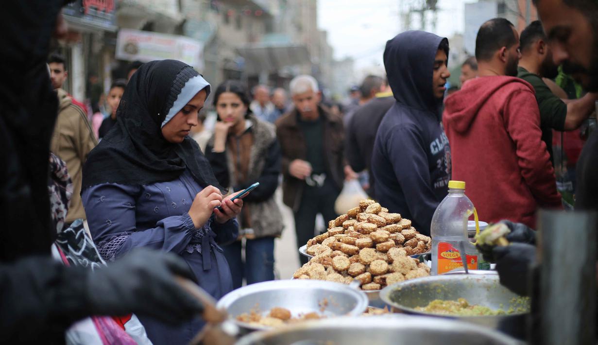 Terlihat upaya warga tetap berupaya menjaga semangat hari kemenangan dengan berbelanja kebutuhan pokok dan persiapan Idul Fitri 1447 Hijriah. Tampak dalam foto, seorang perempuan pengungsi Palestina menggunakan telepon seluler sambil berbelanja di pasar jalanan sekitar kamp pengungsi Nuseirat, Jalur Gaza tengah, pada Rabu 18 Maret 2026. (Eyad Baba/AFP)
