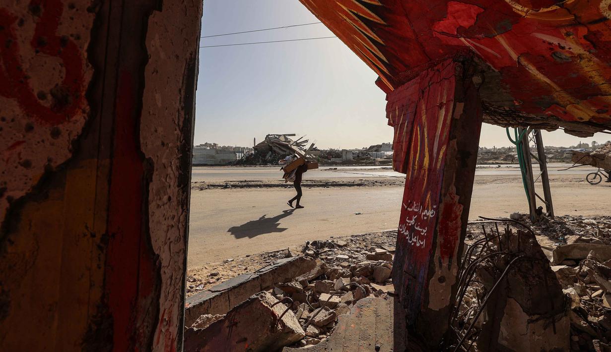 Seruan itu disampaikan Menteri Luar Negeri Mesir, Badr Abdelatty, Sabtu (6/12/2025). Tampak dalam foto, seorang pria membawa kardus berjalan melewati bangunan hancur yang dipenuhi mural di kamp pengungsi Maghazi di luar Deir al-Balah pada Senin 8 Desember 2025. (EYAD BABA/AFP)