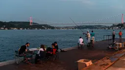 Orang-orang menikmati malam di sebelah pantai Bosphorus ketika jembatan Fatih Sultan Mehmet terlihat di Istanbul (6/7/2020). Ketika diselesaikan pada 1988, jembatan tersebut menjadi jembatan gantung terpanjang ke-5 di dunia; sekarang jembatan tersebut berada pada urutan ke-19. (AFP/Ozan Kose)